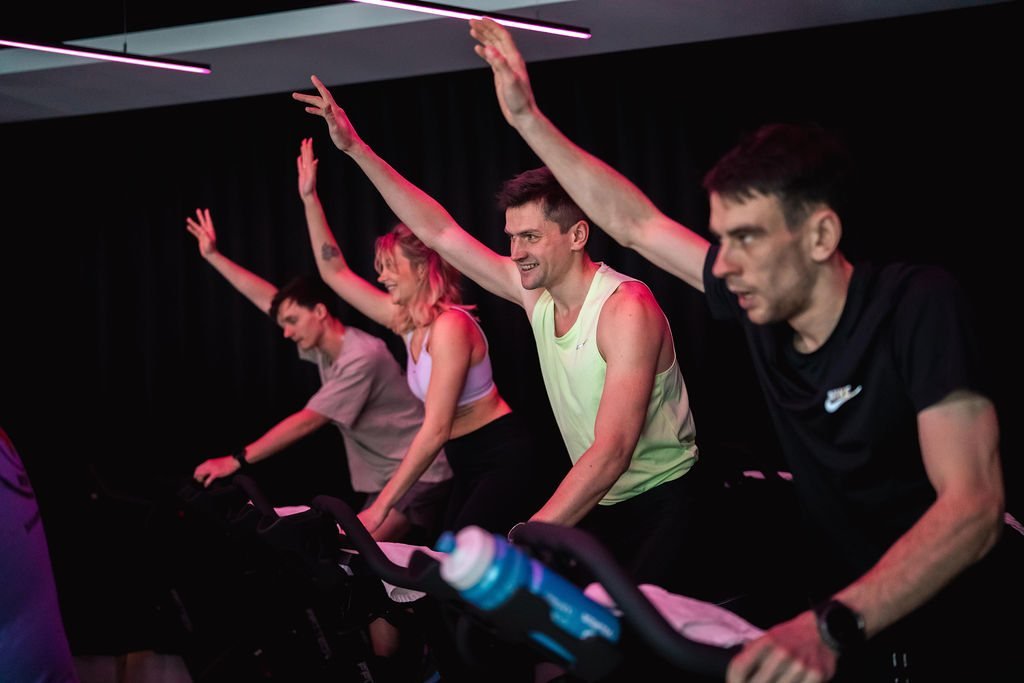 Spin treniruotė Power Fitness studijoje, dalyviai iškėlę rankas pagal muziką